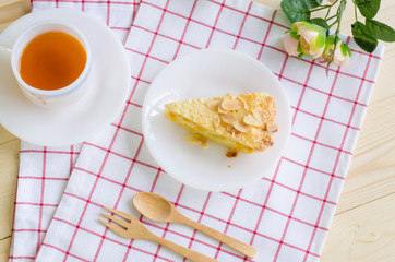 Flat lay image of a piece of delicious corn pie on white ceramic plate over red & white scott pattern cloth with black tea in white cup & saucer, wooden fork & spoon, and small roses over wooden table
