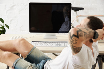 Close focus on relaxing tattoed freelancer dreamily looking somewhere while taking break from work, with his legs on table inside big white loft