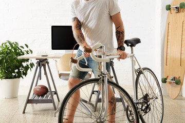 Bearded tattoooed man in blank t-shirt moves his vintage bicycle in big loft in front of his working desktop with computer and printer on it Big wooden longboard and green plant on sides.