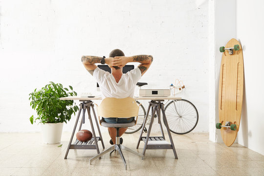 Tattooed man in blank white t-shirt looks in monitor with his hands folded behind head Back view in big loft room with brick wall and longboard, rugby ball, green plant and vintage bicycle around him