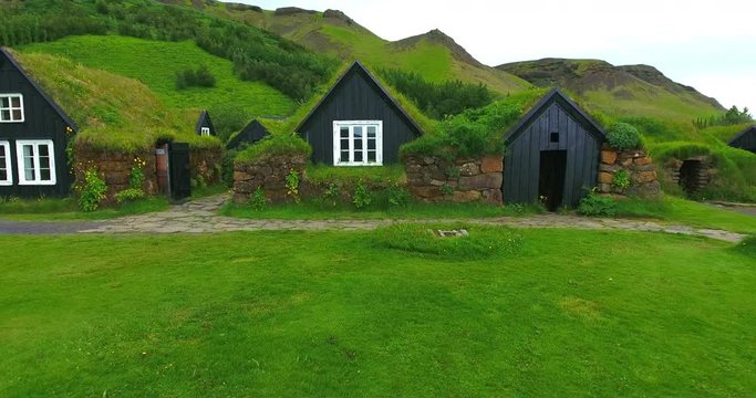 Up close aerial footage of some old turf village houses in Iceland. There is lots of lush greenery, grass, unidentified people waking with a black dog with mountains in the background.