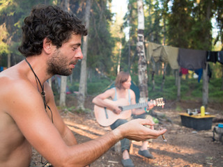 A man in the foreground smokes, and a woman in the background of plays guitar