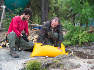 Two men are sitting near a yellow bag on a background of trees