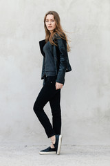 Happy young beautiful woman in black leather jacket black jeans slip-on posing for model tests against textured wall