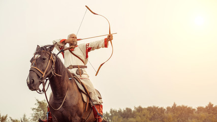 Man in ethnic clothing is riding a horse and aiming from the bow