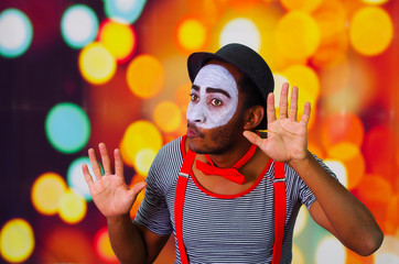 Pantomime man with facial paint posing for camera interacting funny using hands, blurry lights background
