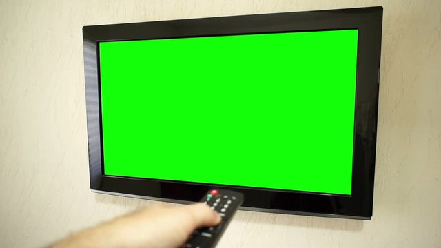 Point of view of an anonymous male hand pressing the remote control pointed at a flatscreen TV and pushing the button to change the channel with a green screen.