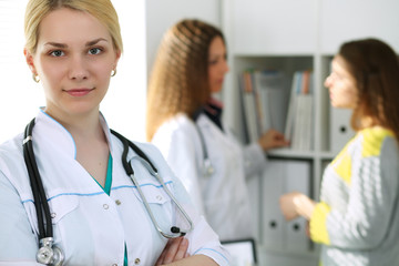 Fototapeta premium Young beautiful female doctor smiling on the background with patient and his physician in hospital. High level and quality medical service concept.