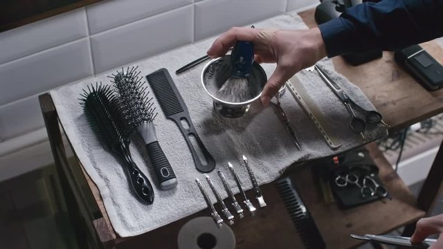 High angle view of barbershop tools on the shelve and male hands taking straight razor and placing bowl with shaving cream and brush in it