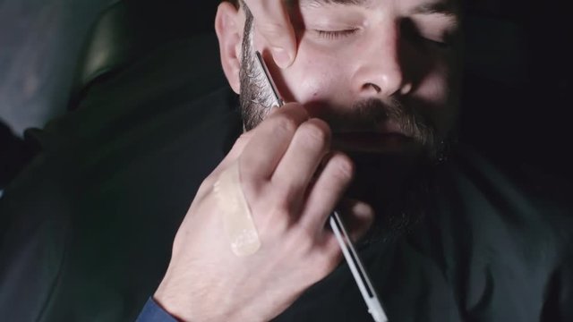 Closeup of hands of barber shaving beard of man with blade straight razor