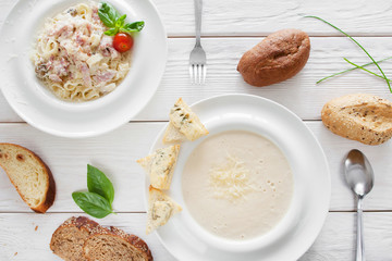 Traditional italian dinner in restaurant, flat lay. Cheese cream-soup and spaghetti carbonara served with bread on white wooden background. European cuisine concept