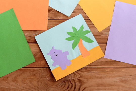 Paper card with happy hippo and a palm tree, colored paper sheets on a wooden table. Kindergarten art and craft activity for kids. Fun children background