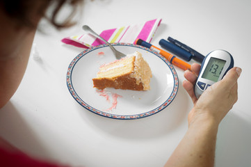 Female hand holding blood sugar meter next to plate of cake. Diabetes, high blood sugar, modern disease concept.