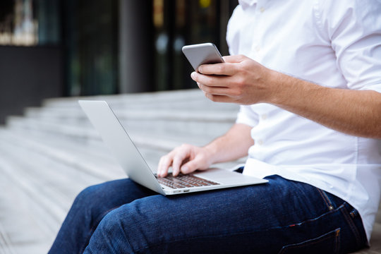 Closeup of young businessman using laptop and cell phone outdoors