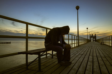 The sad guy in hood, sitting on bench  at wooden pier. Baltic sea in the evening.