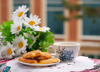Завтрак из сырников с медом и чая на фоне белых цветов на балконе. На заднем плане окна здания. Продукты стоят на вышитой салфетке.