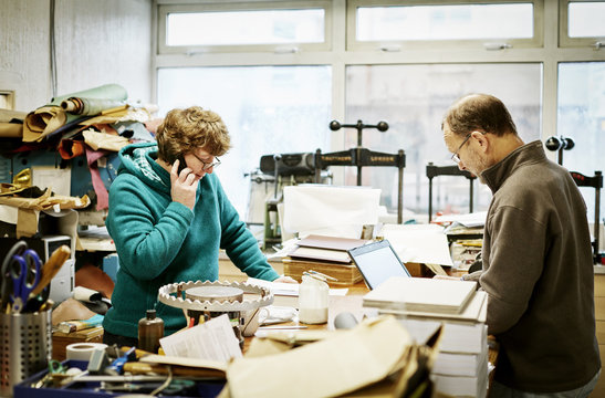A woman taking a phone call and a man working on a laptop computer in a bookbinding workshop. 