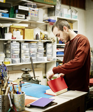 A man working in a book binding workshop, creating a red cover for freshly stitched pages. 