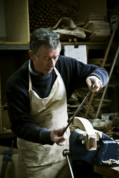 A man working in a furniture maker's workshop using a rasp on a wooden shape in a clamp. 