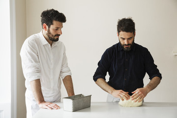 Two bakers standing at a table, kneading bread dough, a metal baking tin.