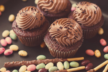 Small chocolate cupcakes and mixture of colorful sweets on black background, retro filter