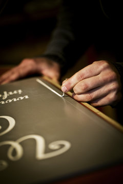 A signwriter working with a loaded brush painting a line freehand on the edge of a sign. 