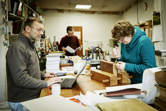 A woman working on the spine of a bound book with a hand tool. A man using a laptop computer. 