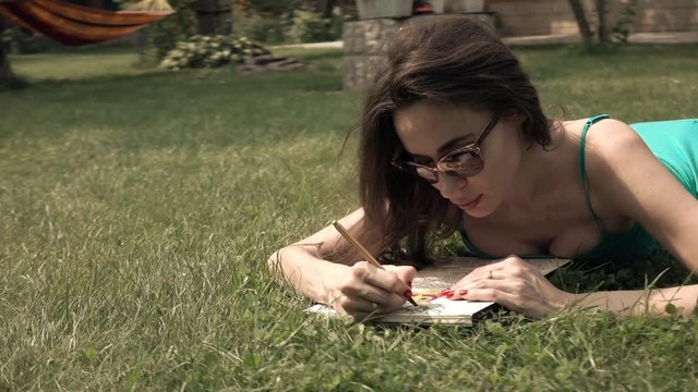 Pretty brunette laying on the lawn and drawing. 4K steadicam wide shot, bleached colors