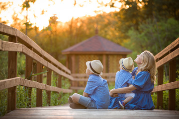 Happy family sitting on the wooden bridge in the park and having fun on sunset outdoors. Mother with kid boy and little toddler boy walking in the park on a sunny day. Smiling woman with children.