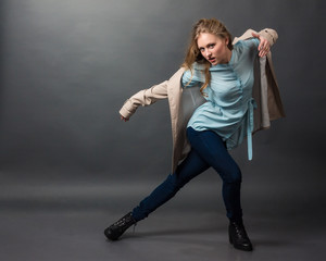 Beautiful blonde full-length in a blue blouse, dark blue jeans and beige coat coming along on a dark gray background.