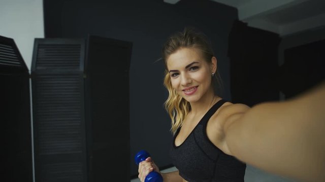 Young blonde woman taking selfie while doing fitness exercises with dumbbells, standing on grey wall background