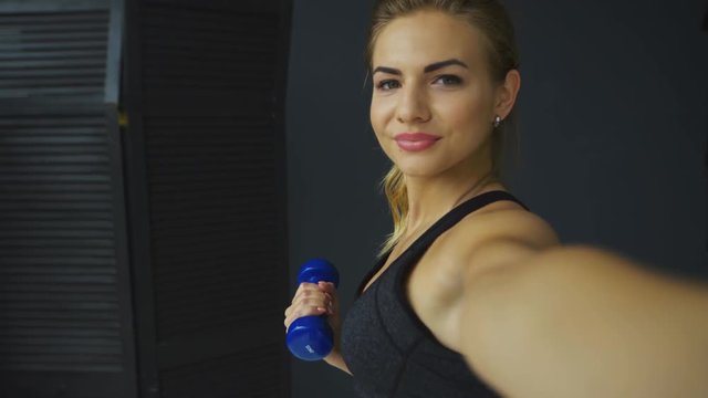 Smiling sporty girl doing dumbbell curls and using app on mobile phone, taking picture, selfie, self-portrait with smartphone