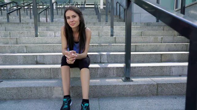 Female urban athlete sitting on stairs after outdoor workout. Sporty woman wearing fashion sport black leggings and blue shirt