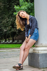 Young cute girl with long hair in a shirt and denim shorts walking in the park in Lviv Striysky sunny summer day posing near the columns at the entrance