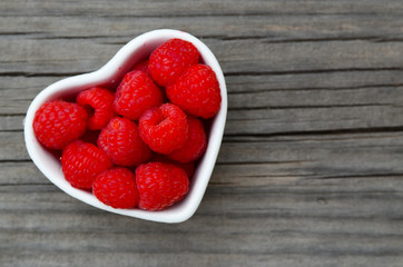 Fresh organic ripe raspberry in a white heart shaped bowl on old wooden background. Selective focus.Healthy food or diet concept.
