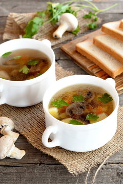 Home mushroom soup in a bowl, wheat bread slices on a cutting board, fresh raw agaricus and green parsley on a wooden table. Vegetable soup with champignons recipe. Closeup