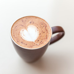 Cup of cappuccino coffee with the milky foam on top in a heart shape viewed from above in a brown cup on white table