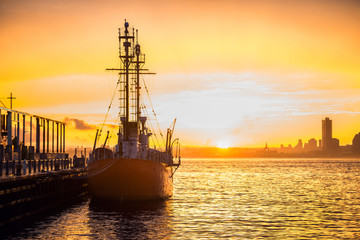 Obraz premium Cargo ship in the harbor at commercial port at sunset time. Sun is going down against beautiful sky at the dock with mooring vessel..