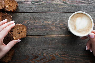 Eating rye bread with coffee, copyspace, wooden background. Top view on female hands holding bread slice and cup of cappuccino, eater pov
