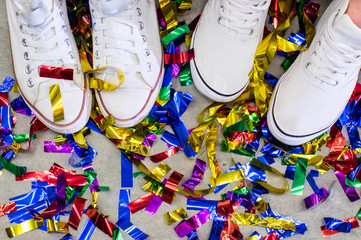 Two feet in white shoes between multicolored festive confetti on the grey floor - 116765565