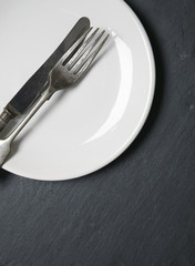 A silver knife and fork with a white plate on a rustic slate background