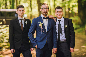 Confident smiling handsome groom in black suit with two groomsman in pine wooden park