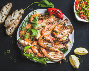 Plate of roasted tiger prawns and octopus with fresh leek, vegetable salad, peppers, lemon, bread and pesto sauce over black background, top view, horizontal composition