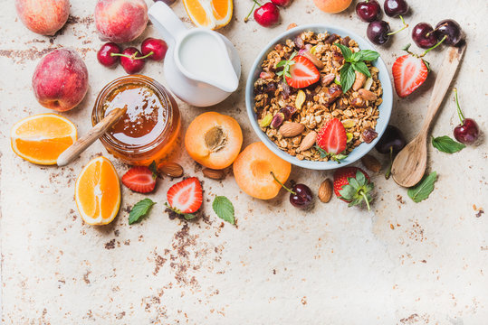 Healthy breakfast ingredients. Oat granola in bowl with nuts, strawberry and mint leaves, milk in pitcher, honey in glass jar, fresh fruits, berries and mint on light concrete background, top view