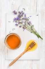 Lavender honey in glass jar served with drizzlier and fresh lavender flowers on ceramic board over white painted wooden backdrop, top view