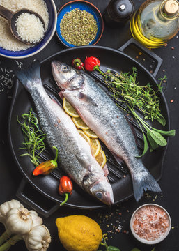 Ingredients for cookig healthy fish dinner. Raw uncooked seabass fish with rice, lemon, herbs and spices on black grilling iron pan over dark background, top view, vertical composition