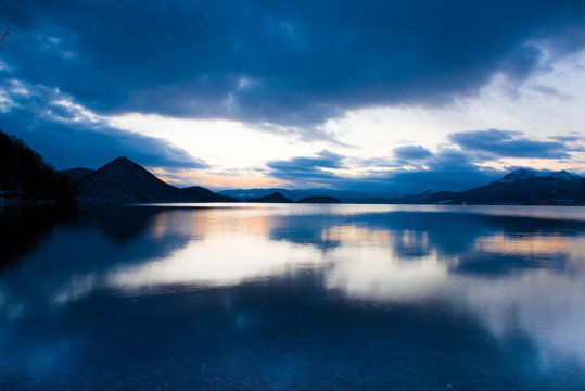 Sunrise over a calm lake with mountains in the background and a semi-cloudy sky.