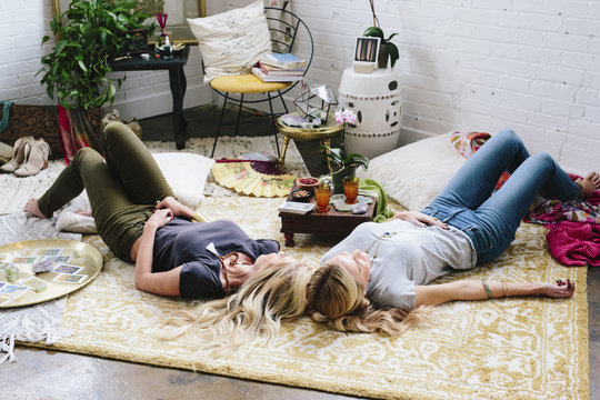 Two women lying on the floor of a room strewn with cushions and personal possessions. 
