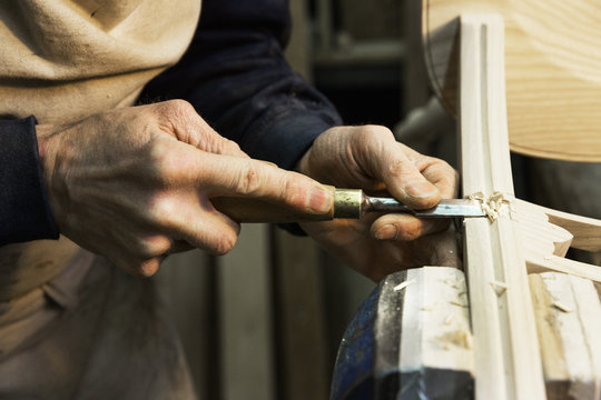 Close up of a man standing at a work bench in a carpentry workshop, working on a wooden chair with a chisel.