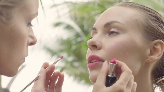 Closeup of model receiving makeup during beach photo shoot, makeup artist adjusting lipstick on her face with brush between palm trees